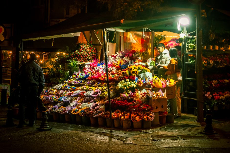 A vibrant flower stall located on Wallington High Street at night, illuminated by bright white lighting. The stall displays an extensive array of fresh floral arrangements, including brightly colored 