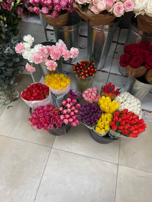 A display of various fresh flower bouquets arranged in metal buckets and wrapped clusters, featuring pink, white, red, yellow, purple, and white carnations and roses. The flowers are set on a tiled fl