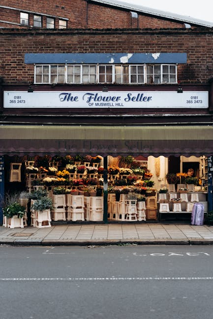 Outside view of The Flower Seller shop on Muswell Hill, displaying a variety of fresh flowers arranged on white shelves and tables beneath a dark awning. The floral displays include colorful blooms li